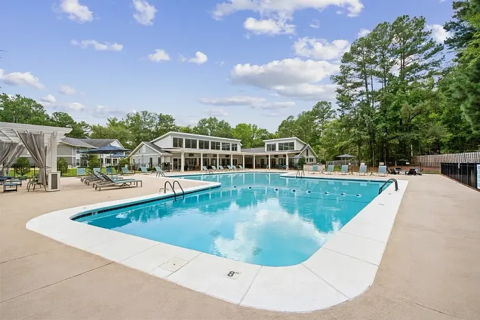 Large outdoor swimming pool with surrounding lounge chairs and umbrellas, set in front of a modern clubhouse and bordered by trees.