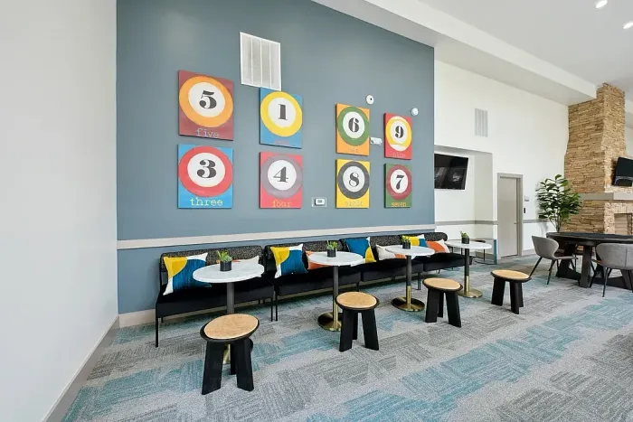 Modern clubhouse seating area with a long bench, small round tables, stools, and colorful numbered wall art, set against a blue accent wall.