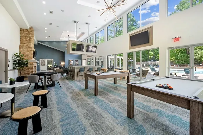Modern clubhouse game room with pool tables, a bar area, seating, large windows, and views of the outdoor pool.