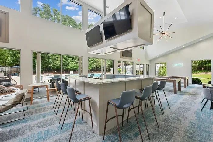 Modern clubhouse bar area with high-top seating, suspended TVs, pool tables, and large windows overlooking the outdoor pool and patio.