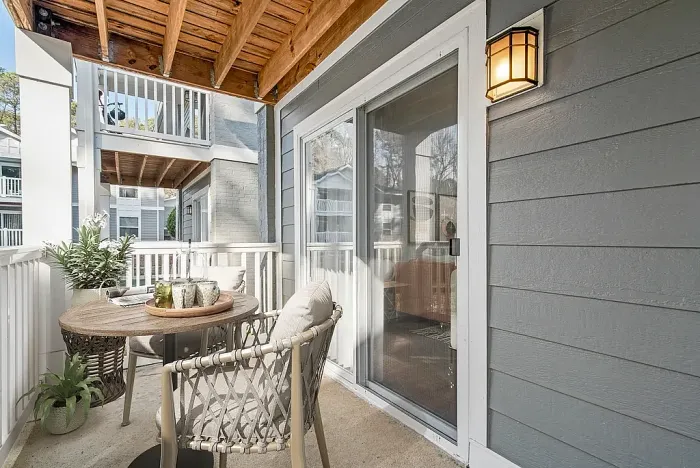 Cozy apartment balcony with a small round table, cushioned chairs, and decorative plants, featuring a sliding glass door, overhead wooden beams, and a view of neighboring units.