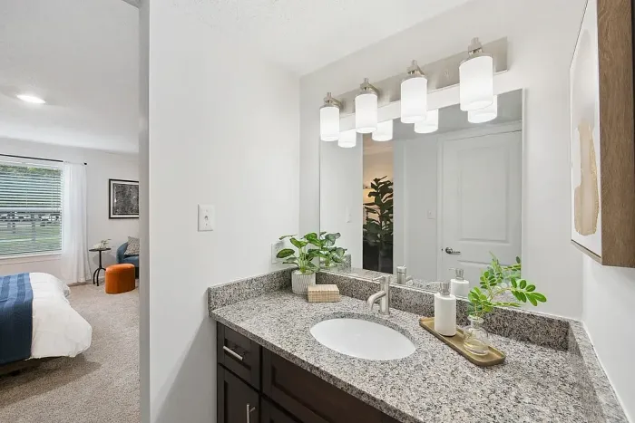 Elegant bathroom with modern lighting and granite countertop adjacent to a serene bedroom with sunlight streaming through large windows.