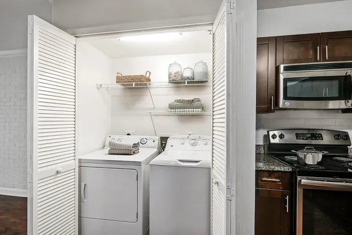 Modern apartment with a compact laundry closet including a washer and dryer, neatly organized with shelves, adjacent to a stylish kitchen featuring stainless steel appliances.
