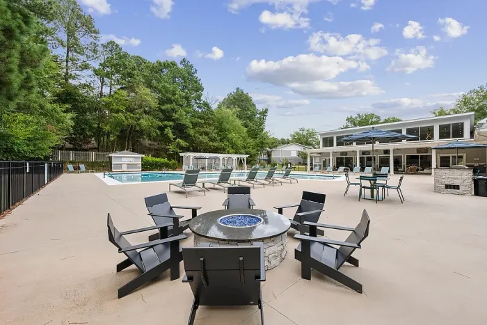 Outdoor community pool area with lounge chairs, umbrellas, and a central fire pit seating area, surrounded by trees and a clubhouse building in the background.