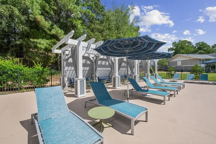 Poolside lounge area with blue chaise chairs, striped umbrellas, and a shaded pergola seating space, surrounded by trees and greenery.