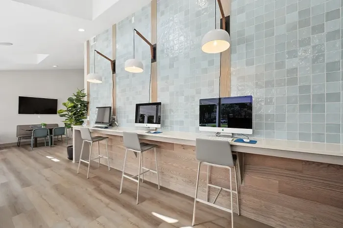 Modern resident business center with a long counter, desktop computers, bar-style seating, pendant lighting, and a tiled accent wall.