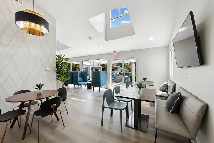 Modern clubhouse lounge with dining tables, booth seating, wall-mounted TV, skylights, and large glass doors opening to an outdoor courtyard.