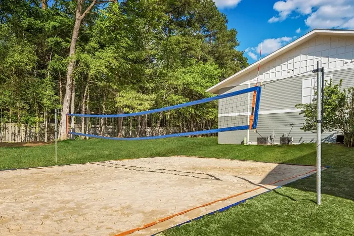 Outdoor sand volleyball court with a net set up beside a residential building, surrounded by grass and trees.