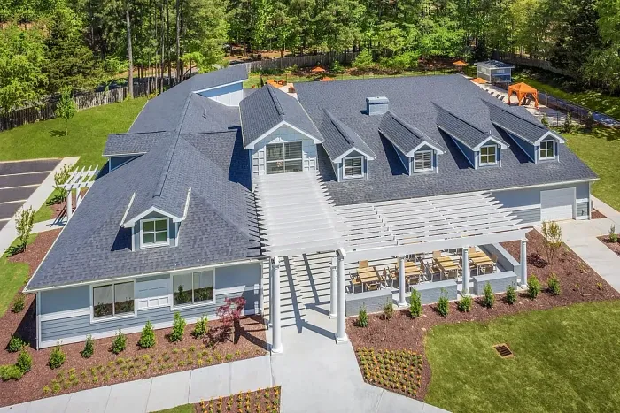 Aerial view of a modern clubhouse with a pitched roof, dormer windows, and a covered outdoor patio with seating, surrounded by landscaped grounds and trees.
