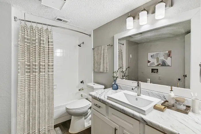 Modern bathroom with a white tub and striped shower curtain, toilet, and a marble-top vanity with a rectangular sink, large mirror, and three-light fixture above. Neutral gray walls, a small plant, and minimal decor create a clean, contemporary look.