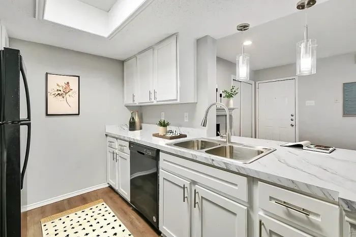 Bright, modern kitchen with white cabinets, marble-look countertops, and a double stainless steel sink with a high-arc faucet. Features include a black refrigerator and dishwasher, pendant lighting, wood-style flooring, and minimal decor with plants and framed wall art.