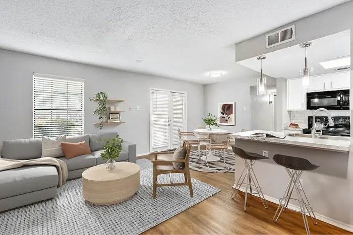Bright open-concept living and dining area with a gray sofa, round coffee table, and accent chair on a textured rug. The space flows into a dining area with a small table and chairs, and a kitchen with a breakfast bar, pendant lighting, and white cabinetry. Large windows and glass doors bring in natural light, complemented by neutral decor and greenery.