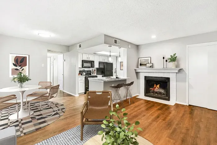 Open-concept living and dining area with hardwood-style flooring, a small dining table and chairs, and a cozy living space featuring a fireplace with a lit fire. The kitchen with white cabinets, black appliances, and a breakfast bar with stools is visible in the background, accented by neutral decor and plants.