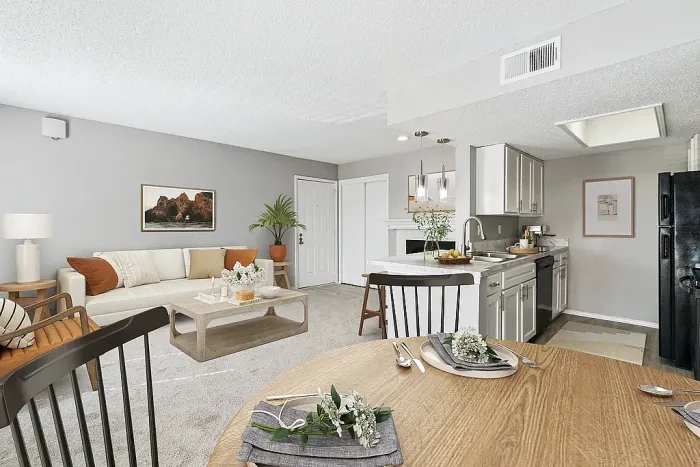 Open-concept living and dining area with plush carpeting, a light-colored sofa, and a coffee table with decor accents. The space connects to a kitchen with white cabinets, marble-style countertops, black appliances, and a breakfast bar with pendant lighting, alongside a dining table set with place settings and greenery, finished with neutral decor and wall art.