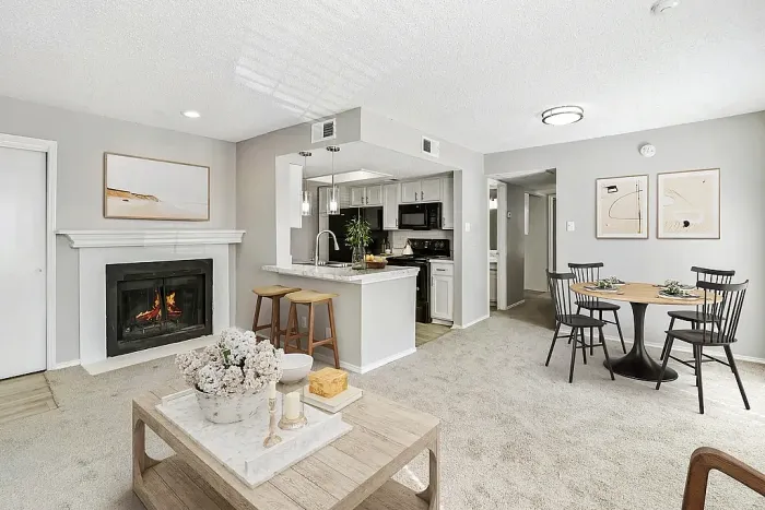Open-concept living and dining area with plush carpet flooring, a cozy fireplace with a lit fire, and a coffee table with decor accents. The space flows into a kitchen with white cabinets, black appliances, and a breakfast bar with stools, alongside a small dining area with a round table and chairs, finished with neutral wall art and decor.