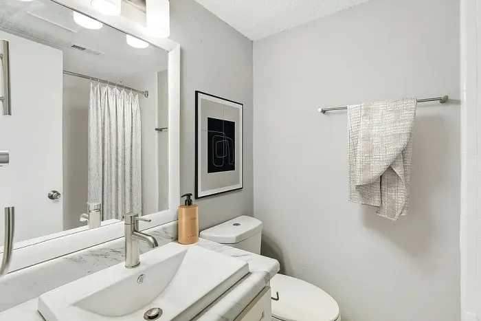 Modern bathroom with a marble-style vanity, rectangular sink, and large mirror with overhead lighting. Features include a toilet, neutral gray walls, a framed abstract print, a towel bar with a textured hand towel, and a white tub with a light shower curtain reflected in the mirror.