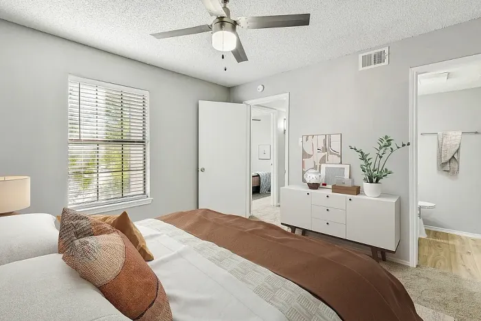 Bright bedroom with plush carpeting, a neatly made bed with neutral bedding and accent pillows, and a ceiling fan with light fixture. Features include a window with blinds, a modern dresser with decor and a plant, and an open doorway leading to an adjoining bathroom.