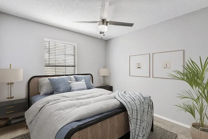 Cozy bedroom with plush carpeting, a neatly made bed with soft gray and blue bedding, and a ceiling fan with light fixture. Features include a window with blinds, nightstands with lamps, framed wall art, and a potted plant for a clean, modern look.