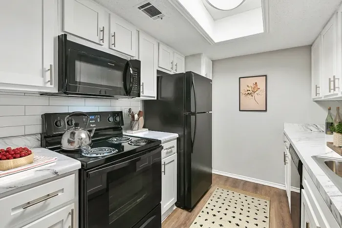 Kitchen with white cabinets, marble-style countertops, and black appliances, including an electric stove, microwave, and refrigerator. Features a subway tile backsplash, wood-style flooring, a small patterned rug, and simple decor with a framed leaf print and countertop accessories.