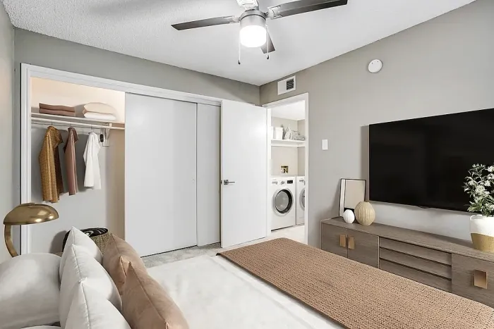 Bedroom with plush carpeting, a neatly made bed with neutral bedding and accent pillows, and a ceiling fan with light fixture. Features include an open closet with shelving and hanging space, a wall-mounted TV with a media console, and an adjoining laundry area with a washer and dryer.
