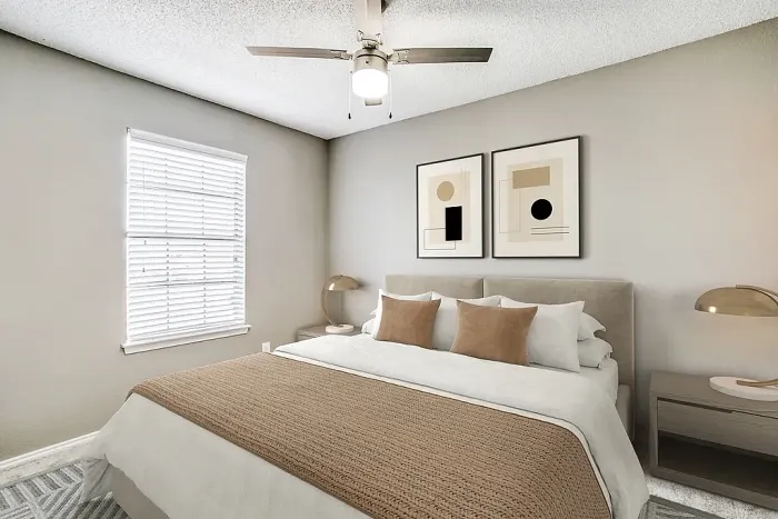Bright bedroom with plush carpeting, a neatly made bed featuring neutral bedding and accent pillows, and a ceiling fan with light fixture. Features include a window with blinds, matching nightstands with modern lamps, and framed abstract wall art for a clean, contemporary look.