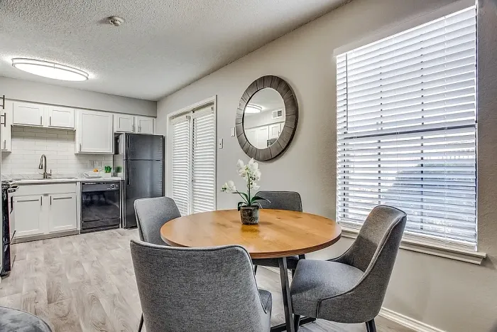 Dining area with hardwood-style flooring, featuring a round wood table and four upholstered chairs. The space connects to a kitchen with white cabinets, marble-style countertops, and black appliances, with large windows, a french door with blinds, and a decorative wall mirror adding natural light and dimension.