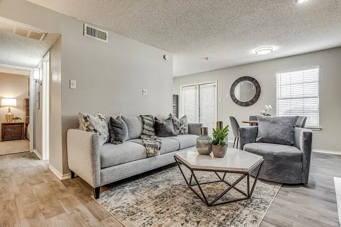 Open-concept living and dining area with hardwood-style flooring, featuring a gray sofa, accent chair, and a coffee table with decor accents on a patterned rug. The space connects to a dining area with a round table and chairs, with large windows, a sliding glass door with blinds, and neutral decor throughout.