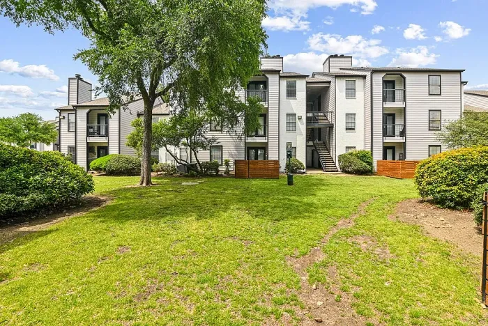 Landscaped apartment community courtyard with a spacious green lawn, mature trees, and walking paths, surrounded by multi-story residential buildings with balconies and exterior staircases. Features include wood privacy fencing, trimmed shrubs, and open outdoor space.