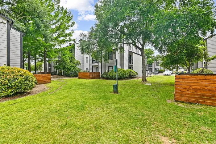 Landscaped apartment community courtyard with green lawn, mature trees, and walking paths, surrounded by residential buildings with balconies. Features include wood privacy fencing, shrubs, and open green space for outdoor enjoyment.