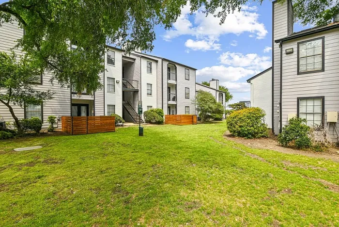 Apartment community courtyard with a green lawn, mature trees, and walking paths, surrounded by multi-story residential buildings with balconies and exterior staircases. Features include wood privacy fencing, landscaped shrubs, and open outdoor space.