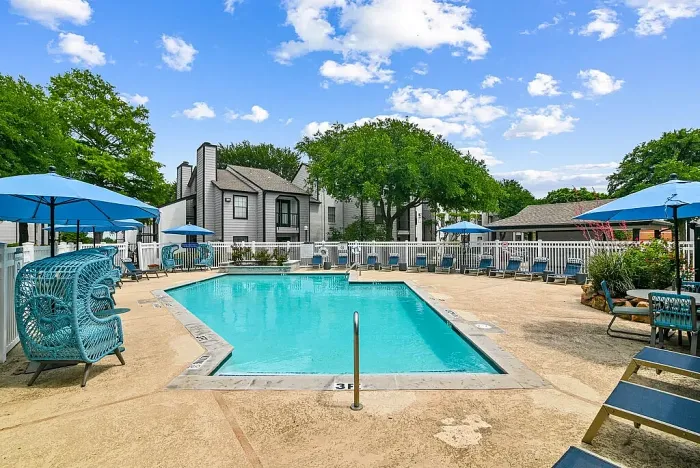 Outdoor community swimming pool with a spacious deck, surrounded by lounge chairs and blue umbrellas. Features include a white fence, shaded seating areas, and landscaped greenery, with residential buildings visible in the background under a bright sky.