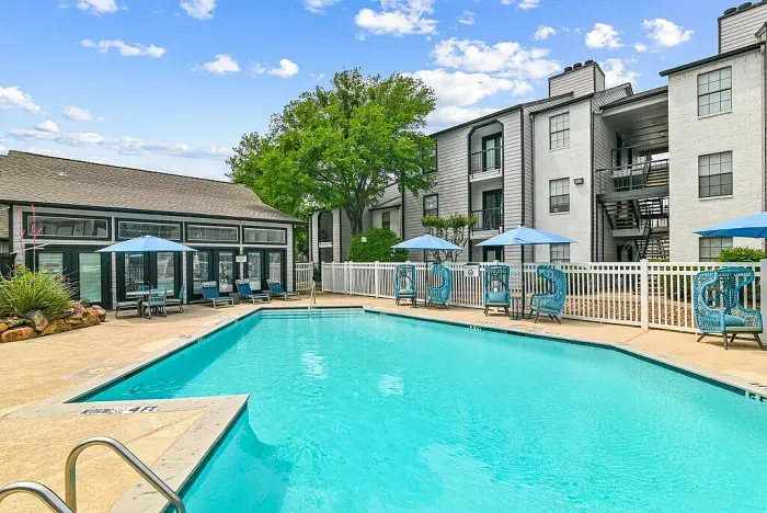Outdoor community swimming pool with a spacious deck, surrounded by lounge chairs, blue umbrellas, and stylish seating. Features include a white fence, a clubhouse building, and landscaped greenery, with apartment buildings in the background under a bright sky.