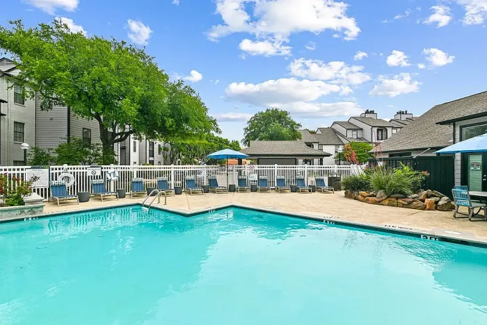 Outdoor community swimming pool with a spacious deck, lined with lounge chairs and blue umbrellas. Features include a white fence, landscaped greenery, and surrounding residential buildings, creating a relaxed outdoor setting under a bright sky.