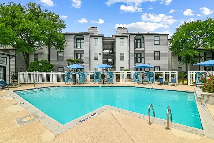 Outdoor community swimming pool with a spacious deck, surrounded by lounge chairs, blue umbrellas, and stylish seating. Features include a white fence, a clubhouse building, and landscaped greenery, with apartment buildings in the background under a bright sky.