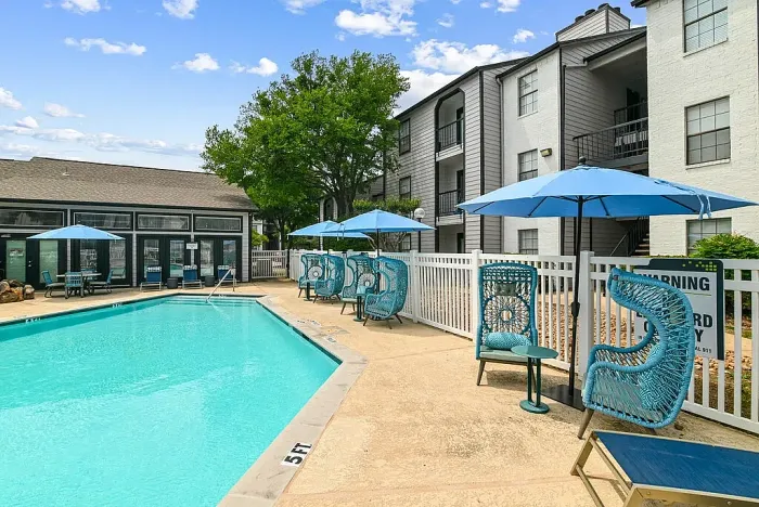Outdoor community swimming pool with a spacious deck, surrounded by lounge chairs, blue umbrellas, and stylish seating. Features include a white fence, a clubhouse building, and landscaped greenery, with apartment buildings in the background under a bright sky.