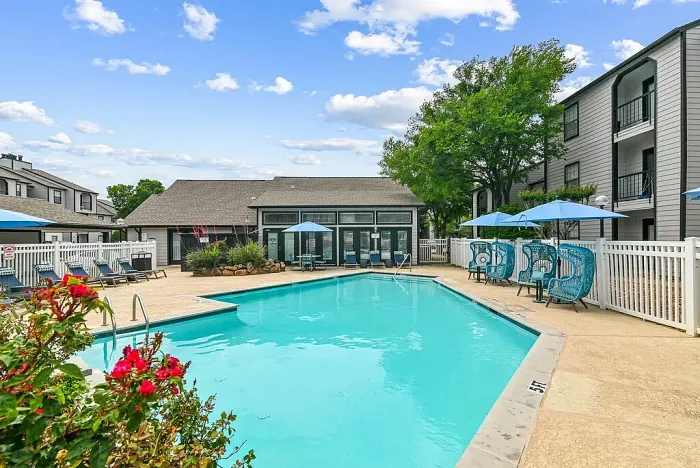 Outdoor community swimming pool with a spacious deck, surrounded by lounge chairs, blue umbrellas, and stylish seating. Features include a white fence, a clubhouse building, and landscaped greenery, with apartment buildings in the background under a bright sky.