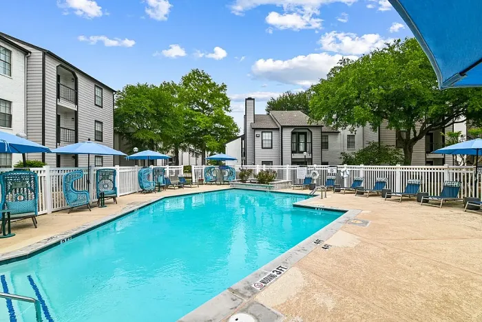 Outdoor community swimming pool with a spacious deck, surrounded by lounge chairs, blue umbrellas, and stylish seating. Features include a white fence, a clubhouse building, and landscaped greenery, with apartment buildings in the background under a bright sky.