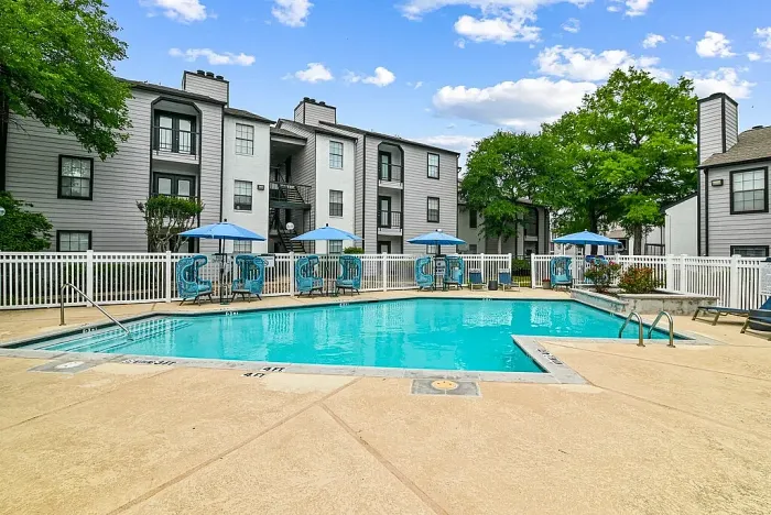 Outdoor community swimming pool with a spacious deck, surrounded by lounge chairs, blue umbrellas, and stylish seating. Features include a white fence, a clubhouse building, and landscaped greenery, with apartment buildings in the background under a bright sky.