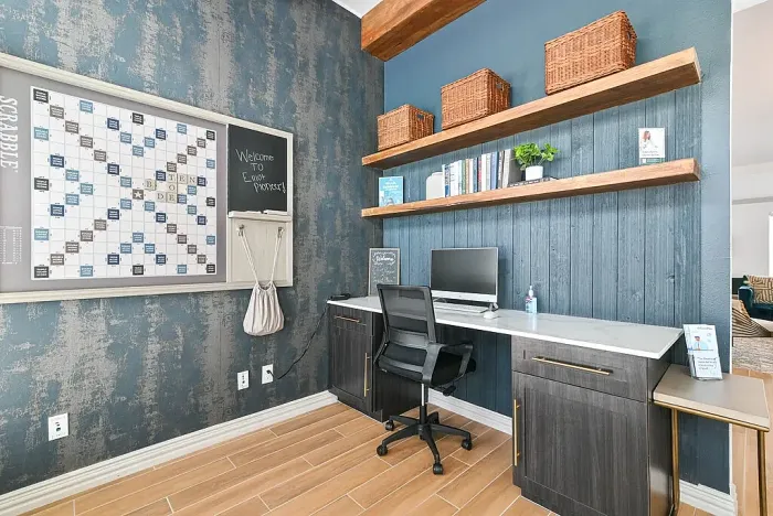 Business center workspace with a built-in desk, computer station, and office chair. Features include wood shelving with books and decor, a marble-style desktop, hardwood-style flooring, and a wall-mounted game board and chalkboard for added community use.