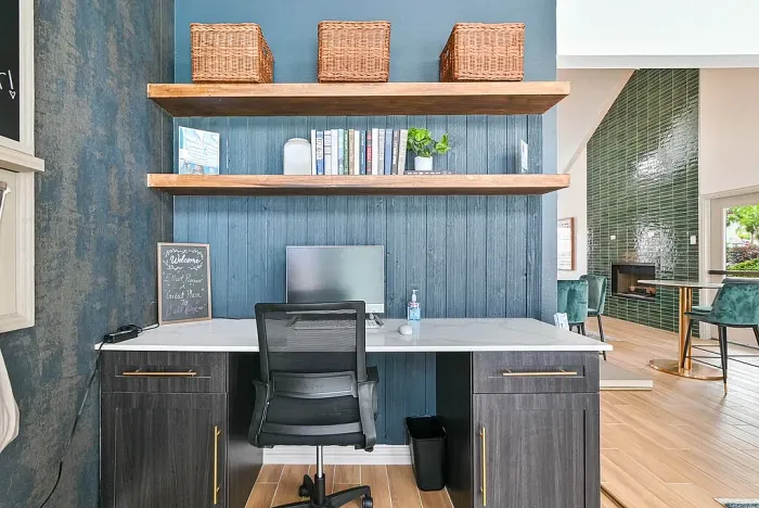 Business center workspace with a built-in desk, computer station, and office chair. Features include wood shelving with books and decor, a marble-style desktop, and hardwood-style flooring, with a modern lounge area and fireplace visible nearby.