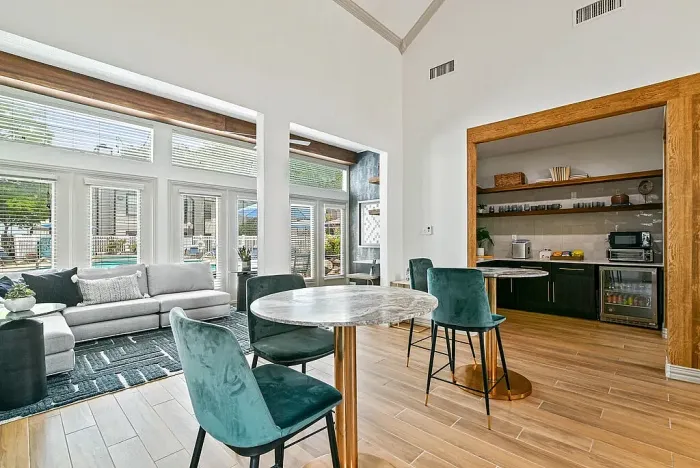 Resident lounge with hardwood-style flooring, featuring round caf&eacute; tables with upholstered chairs and a seating area with a sofa. Includes a kitchenette with shelving, countertop appliances, and a beverage fridge, with large windows providing natural light and views of the pool area.