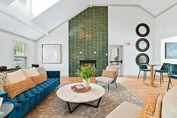 Resident lounge with hardwood-style flooring and a seating area featuring a sofa, accent chairs, and a round coffee table on a patterned rug. Highlights include a fireplace with a green tile accent wall, high ceilings, natural light from large windows, and additional caf&eacute;-style seating.