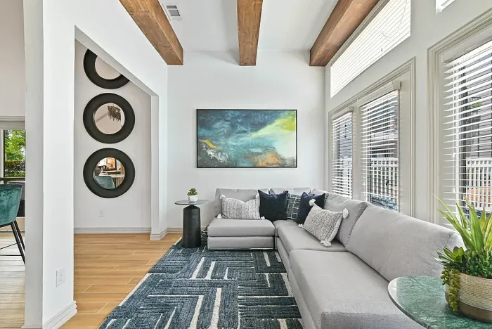 Resident lounge with hardwood-style flooring and a seating area featuring a sectional sofa, accent tables, and a patterned rug. Highlights include exposed wood ceiling beams, large windows with blinds for natural light, and wall art with decorative mirrors.