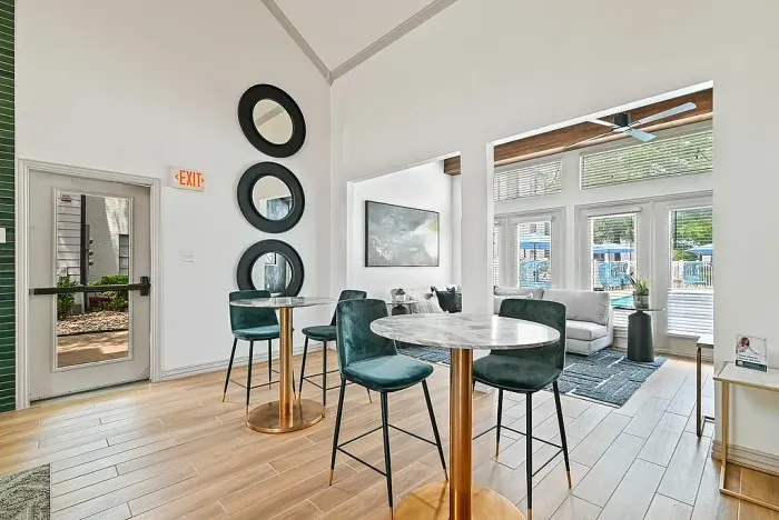 Resident lounge with hardwood-style flooring, featuring caf&eacute;-style tables with upholstered chairs and a bright seating area in the background. Highlights include high ceilings, decorative wall mirrors, large windows for natural light, and an open layout connecting to a comfortable lounge space.