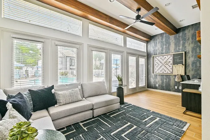 Resident lounge with hardwood-style flooring, featuring a sectional sofa and patterned rug. Highlights include exposed wood ceiling beams, large windows with blinds for natural light and pool views, and a built-in business center with desk.