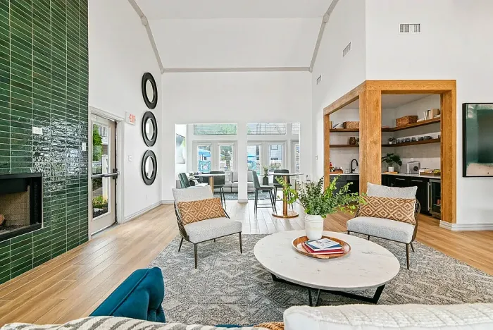 Spacious resident clubhouse with vaulted ceilings, featuring a seating area with accent chairs and coffee table, hardwood-style flooring, a green tile fireplace wall, and a kitchenette with open shelving. Large windows and glass doors bring in natural light and offer views of the outdoor pool area.