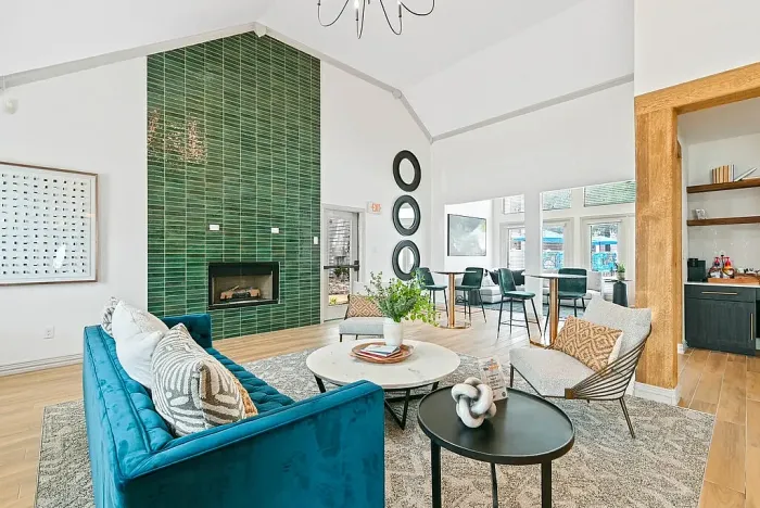Bright clubhouse lounge with vaulted ceilings, featuring a teal sofa, accent chairs, and a coffee table arranged on a patterned rug. A floor-to-ceiling green tile fireplace serves as a focal point, with nearby high-top seating and a kitchenette area. Large windows and glass doors bring in natural light and offer views of the outdoor pool.