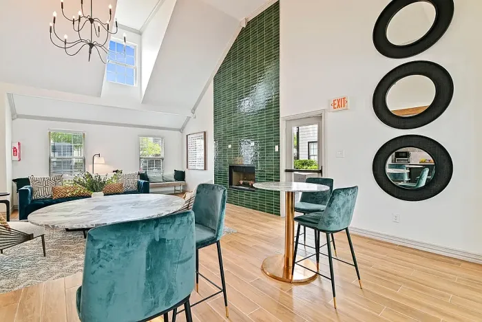 Open clubhouse space with high ceilings and a modern chandelier, featuring round tables with upholstered chairs, hardwood-style flooring, and a floor-to-ceiling green tile fireplace. A cozy seating area with a sofa is visible in the background, along with decorative wall mirrors and natural light from surrounding windows.