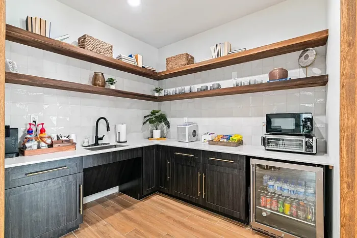 Modern clubhouse kitchenette with dark cabinetry, white countertops, and open wood shelving. Features include a sink, microwave, toaster oven, mini fridge stocked with drinks, and a coffee/snack station, with decorative baskets, books, and plants displayed on the shelves.