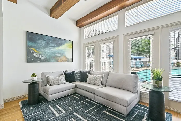 Cozy clubhouse seating area with a gray sectional sofa, accent pillows, and a patterned area rug. Floor-to-ceiling windows with blinds provide natural light and views of the outdoor pool, while wood ceiling beams and wall art add a modern touch.